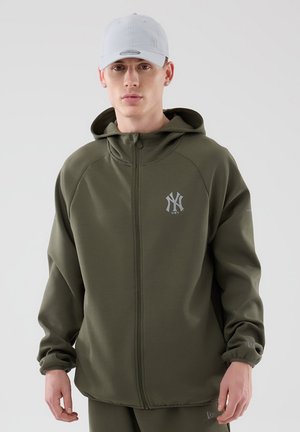 Joven con sudadera con cremallera de los New York Yankees de color verde oliva y gorra de béisbol de los Yankees de color gris claro, posando frente a un fondo liso.