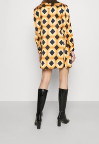 Robe à motif en diamants jaune et noir avec des manches longues et des boutons, associée à des bottes noires en cuir brillant jusqu'au genou avec des talons.