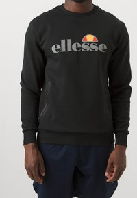 Sweat-shirt noir à col rond et manches longues, arborant un grand logo "ellesse" gris et un emblème multicolore. Deux poches zippées.