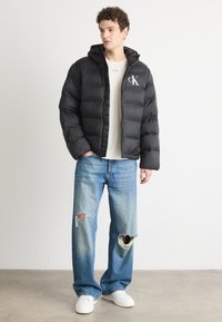 Schwarze Pufferjacke mit Kapuze und CK-Logo, strukturiertem Stoff, kombiniert mit hellblauen, zerrissenen Jeans und weißen Turnschuhen.