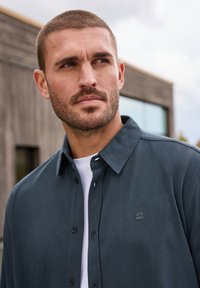 Mörkblå, knappad skjorta med krage och en liten logotyp på bröstet. Har en slät textur och en subtil glans på tyget.