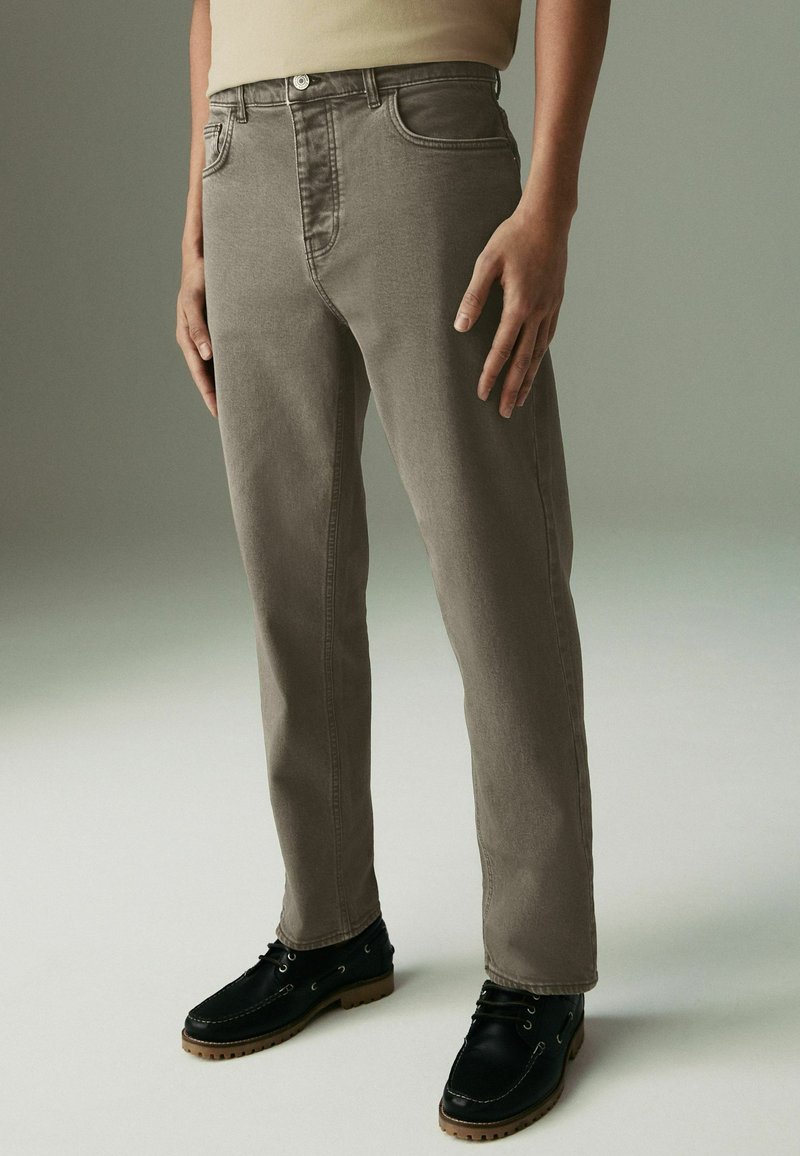 Jeans in denim grigio con una vestibilità raffinata, dotati di cinque tasche, chiusura con zip e una finitura leggermente scolorita, abbinati a scarpe nero con lacci.