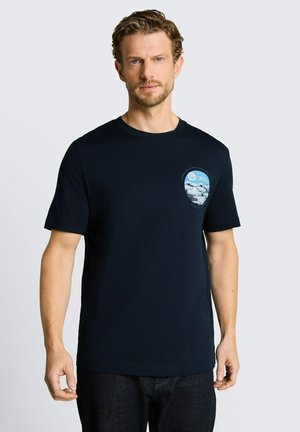 Mężczyzna z krótkimi kręconymi włosami i brodą, ubrany w czarny t-shirt z małą niebieską grafiką oceanu na lewej piersi.