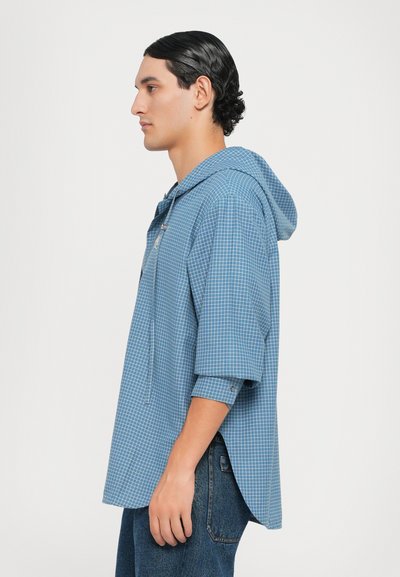 Camisa con capucha de gran tamaño a cuadros azules, con mangas de tres cuartos y un detalle de cremallera en la parte delantera, hecha de tejido ligero.