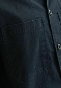 Donkerblauwe shirt met een gestructureerd ontwerp, voorzien van een borstzak en ronde zwarte knopen. Gelaagd stof met zichtbare stikseldetails.