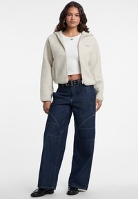 Felpa con zip color crema abbinata a pantaloni in denim scuri a gamba larga con dettagli cuciti e una cintura nera con accessori in argento.