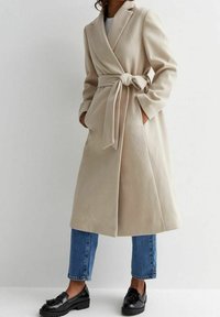 Abrigo de lana beige con cinturón, mangas largas, cuello muesca y bolsillos laterales, combinado con jeans de pierna recta azul y mocasines negros.
