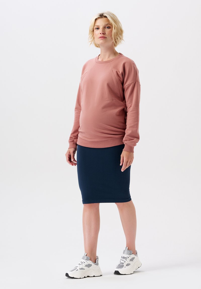 Femme blonde portant un sweat-shirt rose poussiéreux, une jupe à côtes bleu marine et des baskets blanches épaisses, se tenant contre un fond blanc uni.
