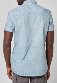 Mies, joka pitää yllään vaaleansinistä lyhythihaista paitaa ja harmaat ruudulliset cargo-shortsit, nähtynä takaa neutraalia taustaa vasten.