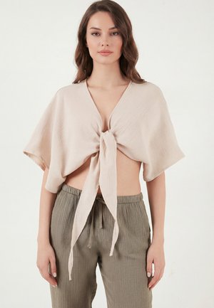 Kobieta ubrana w beżowy krótki top z krótkim rękawem i wiązaniem z przodu oraz luźne, teksturowane oliwkowozielone spodnie na jednolitym białym tle.