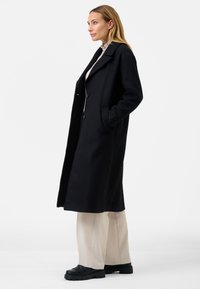 Cappotto nero in lana con colletto a punta, bottoni frontali e tasche laterali, abbinato a pantaloni beige a gamba larga e scarpe platform nere.