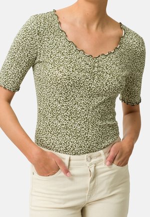 Zelený vzorovaný top s krátkým rukávem s vlnitým výstřihem a lemováním rukávů, nošený s světle béžovými kalhotami, ruce v předních kapsách.