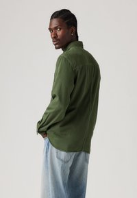 Chemise à manches longues vert olive avec un col boutonné. Tissu légèrement texturé, ourlet légèrement arrondi. Associée à un pantalon en denim clair.