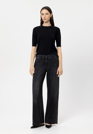 Schwarzes Kurzarmoberteil kombiniert mit weit geschnittenen schwarzen Jeans, die verziertes Seitenornament haben, und spitzen schwarzen Schuhen.