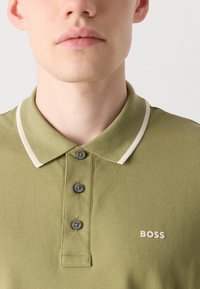 Zelena polo majica iz teksturiranega blaga, s kragnico, tremi gumbi in kontrastnim belim obrobo. Logo "BOSS" v svetli barvi na prsih.