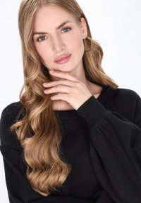 Schwarzer Pullover mit gepufften Ärmeln; glatte, weiche Textur. Das Model hat lange, wellige blonde Haare und trägt schlichte Creolen. Neutraler Hintergrund.