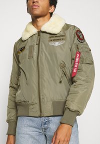 Olivgrüne Bomberjacke mit einem weißen Pelzkragen, Frontreißverschluss, Aufnähern auf der Brust und dem Ärmel sowie gerippten Bündchen und Saum.