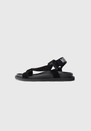 Sort flad sandal med justerbare remme omkring ankel og tå, med hvid tekst på ankelremmen mod en enkel lys baggrund.