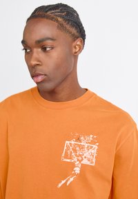Orange bomulds T-shirt med rund halsudskæring, prydet med et hvidt basketballgrafik på brystet. Glat tekstur og afslappet pasform.