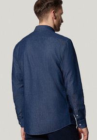 Ciemnoniebieska koszula denimowa z karczkiem z tyłu, długimi rękawami, guzikami przy mankietach oraz zaokrąglonym dołem. Tkanina ma subtelną teksturę i lekki połysk.