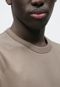 Κοντομάνικο βαμβακερό T-shirt σε γκριζωπή απόχρωση με στρογγυλή λαιμόκοψη και ορατές ραφές. Ομαλή υφή, ύφασμα μεσαίου βάρους.