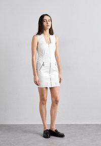 Balta bez piedurknēm mini kleita ar priekšējo rāvējslēdzēju, divām rāvējslēdzēju sānu kabatām un ribotu apdari. Modelis valkā melnas slip-on kurpes.