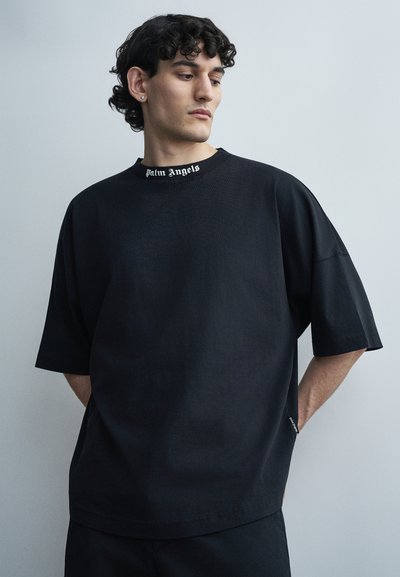 Homme aux cheveux bouclés portant un T-shirt noir surdimensionné avec le texte "Palm Angels" sur le col, debout devant un fond clair uni.