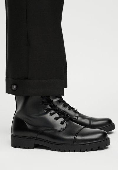 Μαύρα γυαλιστερά δερμάτινα μποτάκια με κορδόνια, φορεμένα με μαύρο παντελόνι με κουμπιά στο τελείωμα, σε ανοιχτό φόντο.