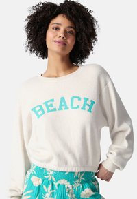 Sudadera de forro polar blanca con "BEACH" en texto turquesa, con un escote ancho y mangas sueltas, combinada con una falda de patrón floral en color verde azulado.