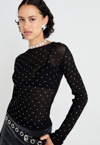 Schwarzes Langarmshirt mit gerippter Textur und verstreuten silbernen Verzierungen. Kombiniert mit einem glänzenden schwarzen Rock und einem dekorativen Gürtel.