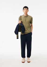 Polo shirt verde oliva con un piccolo logo, abbinato a pantaloni della tuta blu navy con strisce gialle ai lati e sneakers beige. Borsa marrone a spalla.