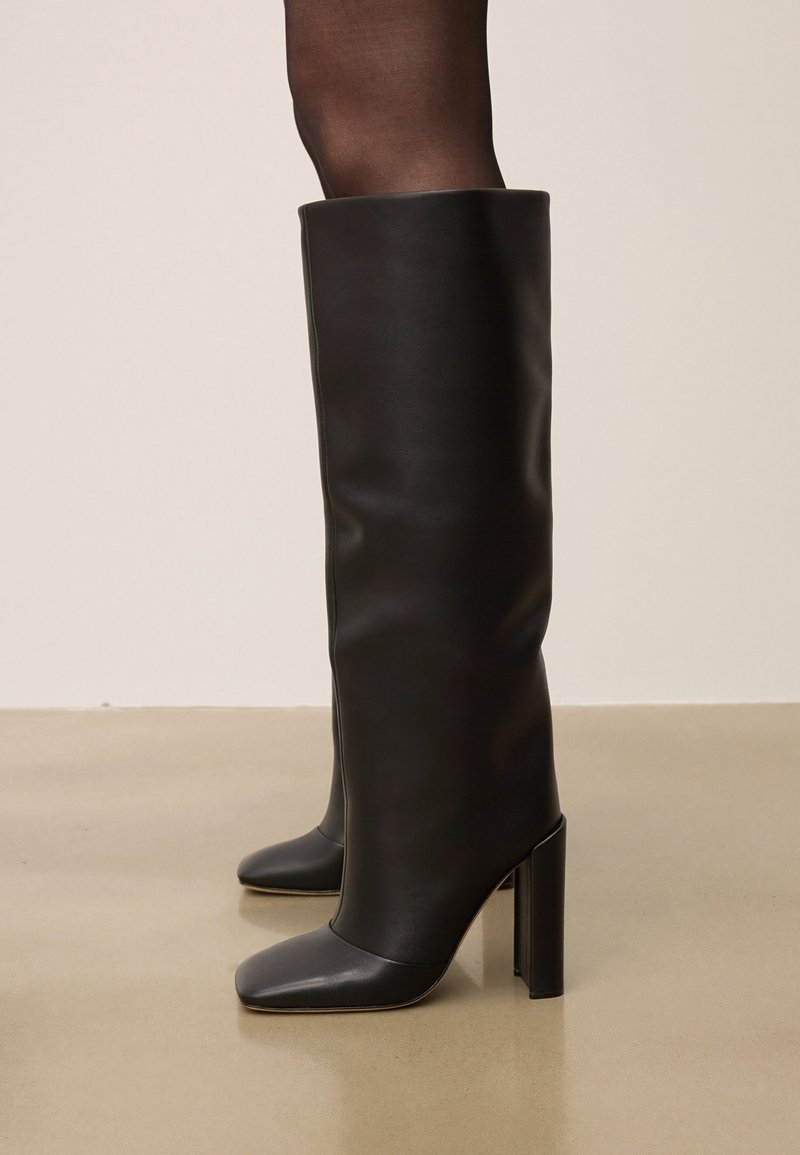 Tall black leather boots with block heels worn over sheer black tights, standing on a smooth beige floor against a plain wall.