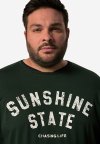 T-shirt di cotone verde scuro con testo bianco audace e consumato che recita "SUNSHINE STATE", con colletto a girocollo in costina.