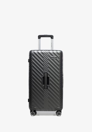 Czarna walizka z twardą skorupą z diagonalnymi rowkami, wysuwanym uchwytem, centralnym uchwytem oraz czterema obrotowymi kółkami, stojąca pionowo na białym tle.