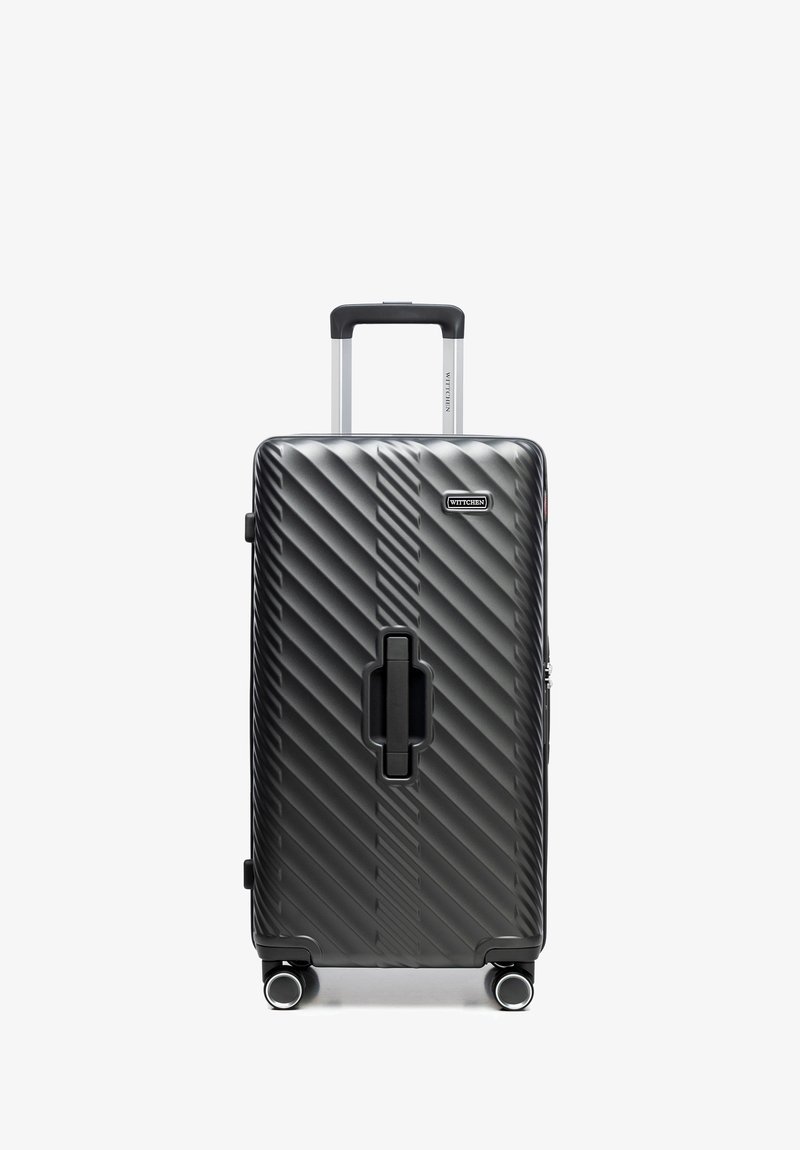 Valigia rigida nera con scanalature diagonali, manico retrattile, impugnatura centrale e quattro ruote girevoli, in posizione verticale su uno sfondo bianco.