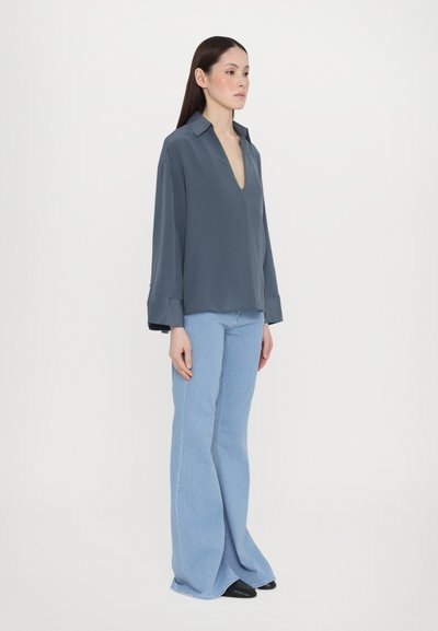 Blusa grigia a maniche lunghe con scollatura a V profonda e polsini ampi, abbinata a jeans a gamba larga blu chiaro e scarpe nere. Design semplice e moderno.