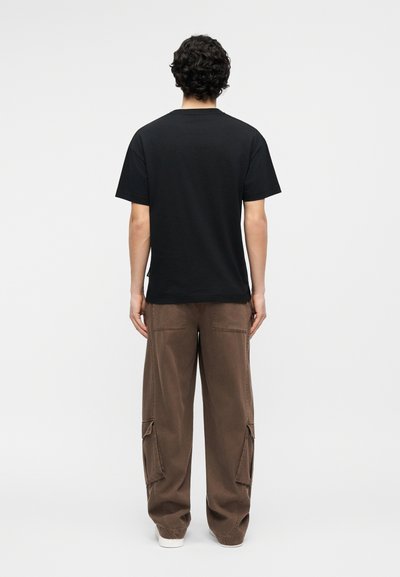 Άντρας με σγουρά μαύρα μαλλιά, φορώντας απλό μαύρο κοντομάνικο μπλουζάκι και άνετο καφέ παντελόνι cargo, στέκεται γυρισμένος πλάτη σε λευκό φόντο.