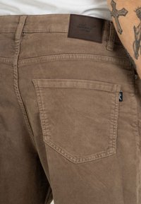 Brown corduroy pants featuring a textured finish, back pocket with a stitched edge, and a leather brand patch at the waistband.