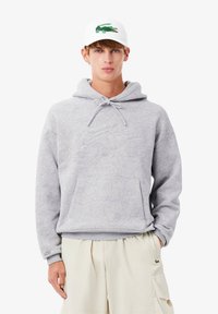 Maglione grigio chiaro con un motivo di delicati disegni floreali, dotato di una tasca a marsupio frontale e cappuccio regolabile con cordino. Abbinato a pantaloni corti beige.