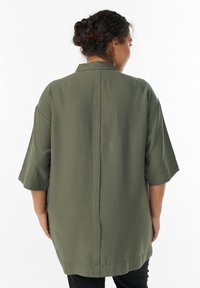 Frau mit geflochtenem Dutt, die ein übergroßes olivgrünes Hemd mit Knöpfen und Ellbogenlangen Ärmeln trägt, mit dem Rücken zur Kamera auf einem weißen Hintergrund.