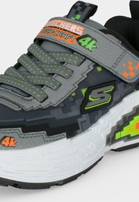 Baskets pour enfants gris et noir avec des touches de vert et d'orange, design pixellisé, logo "Skechers" et bride Velcro étiquetée "Mega Craft 4k."