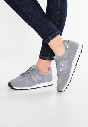 Person iført grå New Balance sneakers og mørkeblå jeans, står med den ene fod let hævet på en hvid gulv.