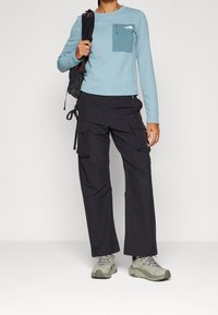 Haut bleu clair à manches longues avec une poche sur le devant ; pantalon cargo noir avec des poches latérales et taille cintrée ; chaussures de randonnée grises.