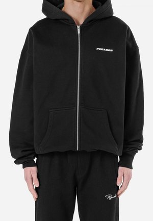 Mann trägt schwarzen Kapuzenpullover mit Reißverschluss und Vordertaschen sowie passende schwarze Hose, beide mit weißen "Pegador"-Logos.