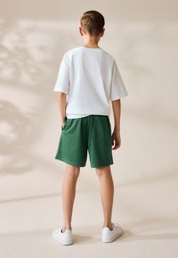 Weißes T-Shirt mit kurzen Ärmeln, kombiniert mit grünen Mesh-Shorts mit seitlichen Taschen. Das Model trägt weiße Turnschuhe. Neutraler Hintergrund.