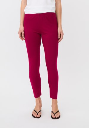 Femme portant des leggings moulants rose foncé, un haut blanc et des sandales noires à bride, debout devant un fond clair uni.