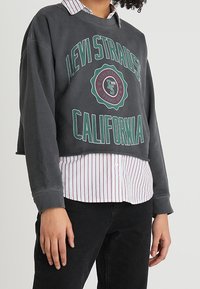 Sudadera gris recortada con texto gráfico verde "Levi Strauss California", superpuesta sobre una camisa de botones blanca y roja a rayas. Pantalones negros visibles.