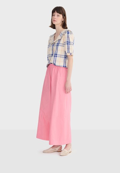 Femme portant un chemisier à carreaux beige et bleu avec un col à volants, un pantalon large rose et des chaussures beige à enfiler, debout de profil.