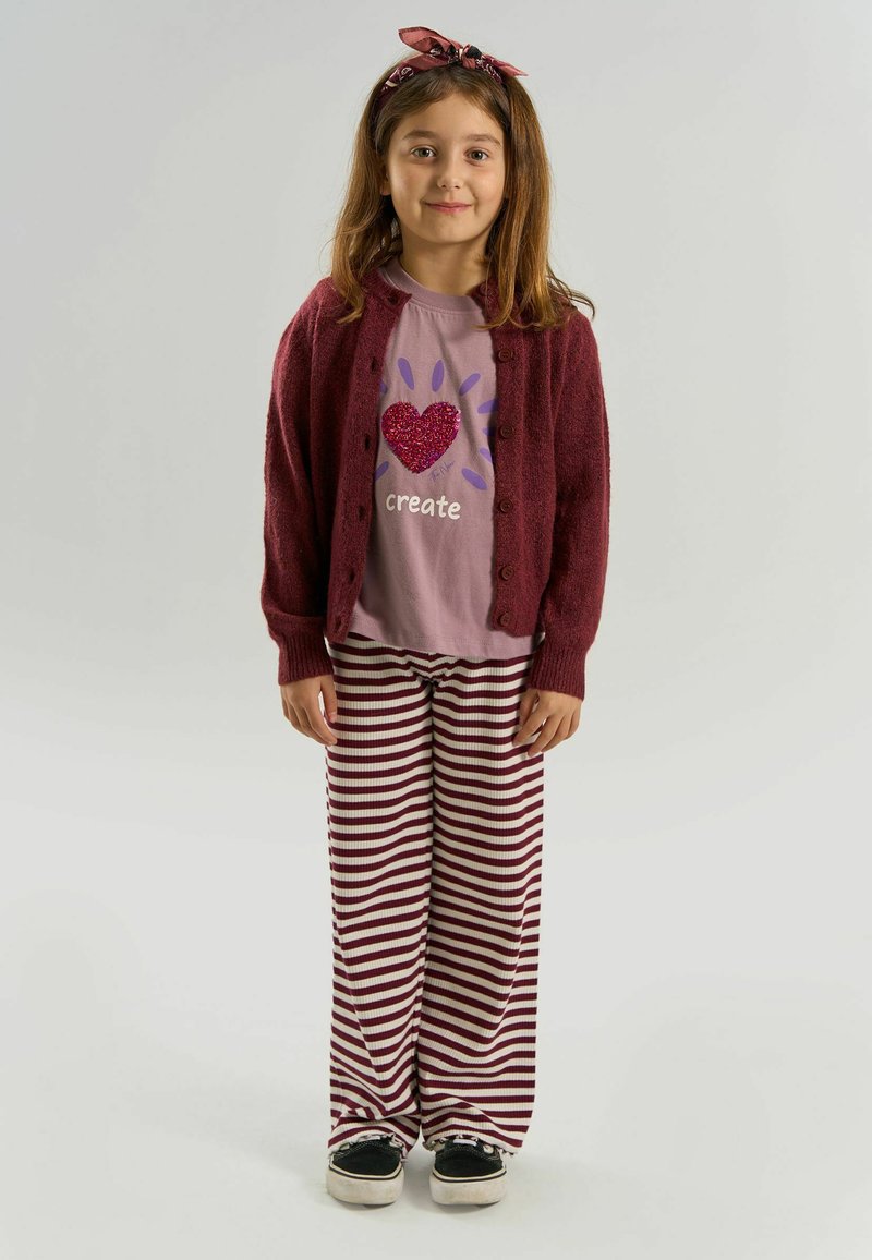 Jeune fille debout, portant un t-shirt rose pailleté avec un cœur "create", un cardigan marron, un pantalon rayé, des baskets noires et un serre-tête marron avec un nœud.