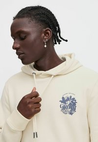 Hoodie de couleur crème en tissu doux avec un graphique en façade mettant en avant un design floral bleu et le texte "GRANDIS AU FUR ET À MESURE". Cordons de serrage ajustables.